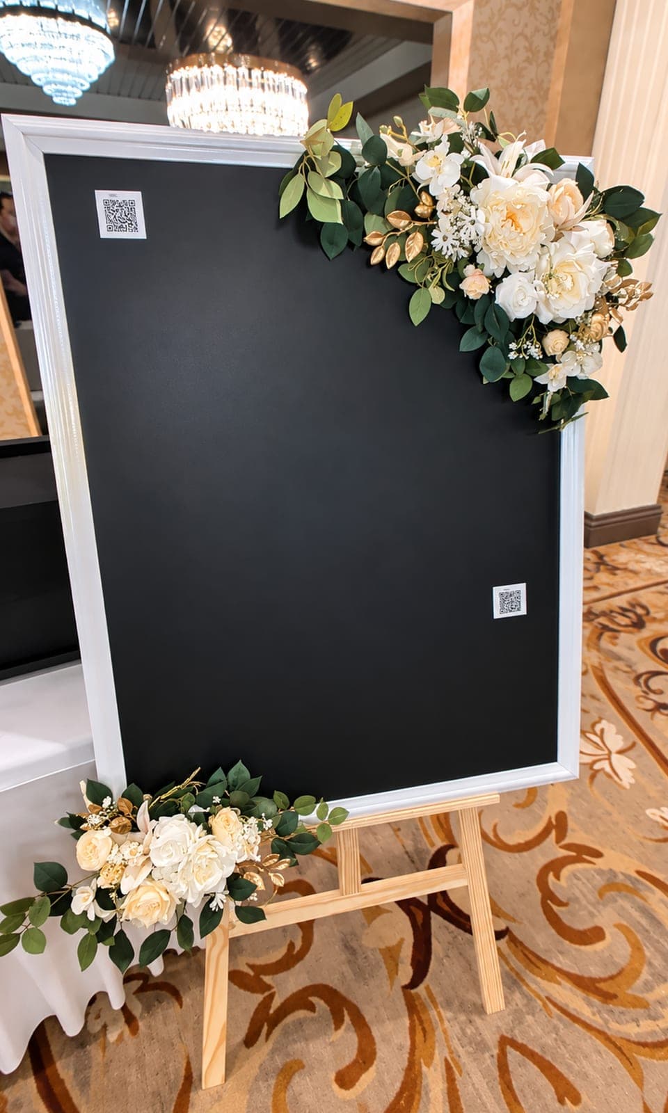 The Tender Kept Memory Board on an easel with white floral accents and QR codes before guests arrive